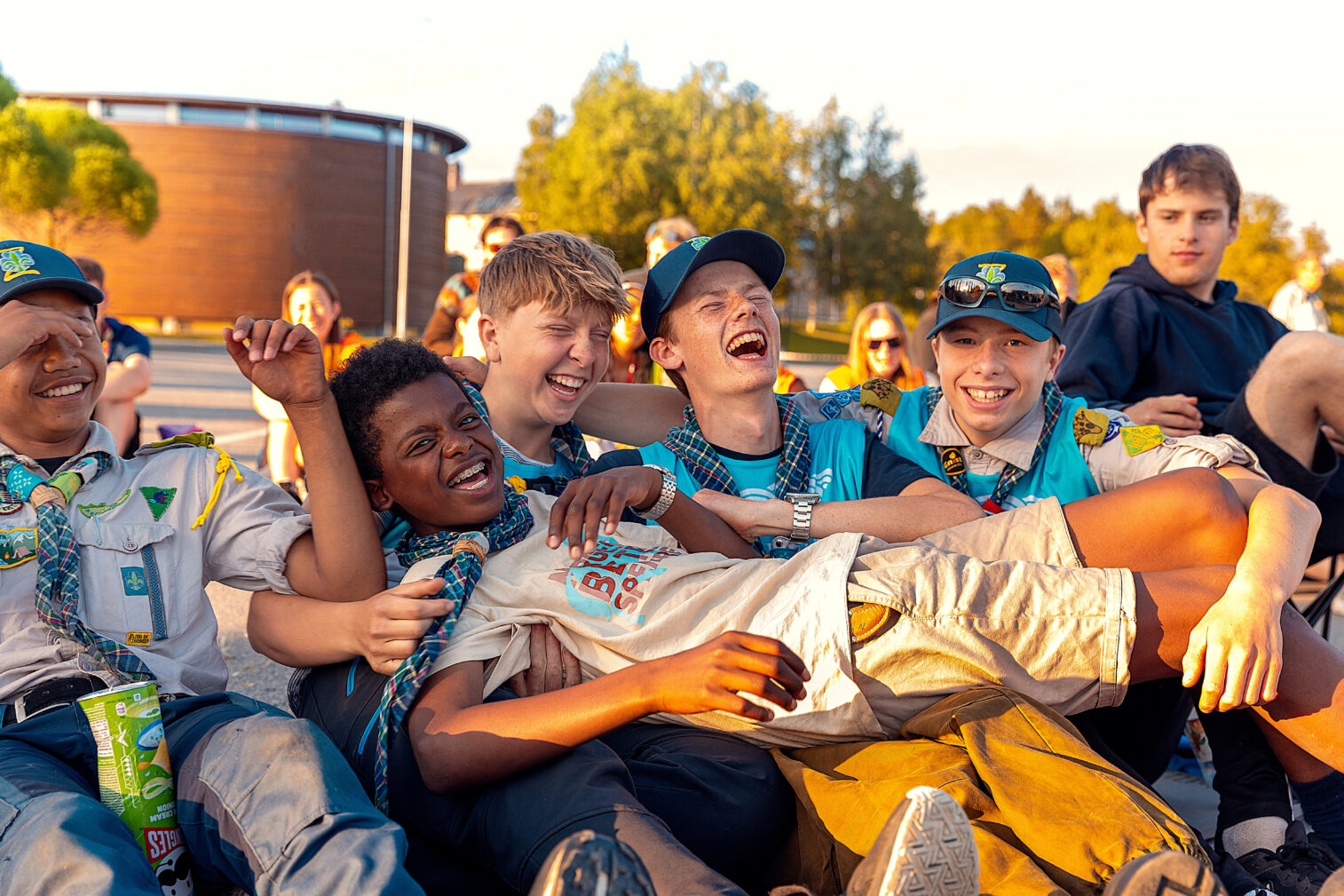 En gruppe leende unge speidere i uniform sitter sammen utendørs og nyter et morsomt øyeblikk i gyllent sollys.