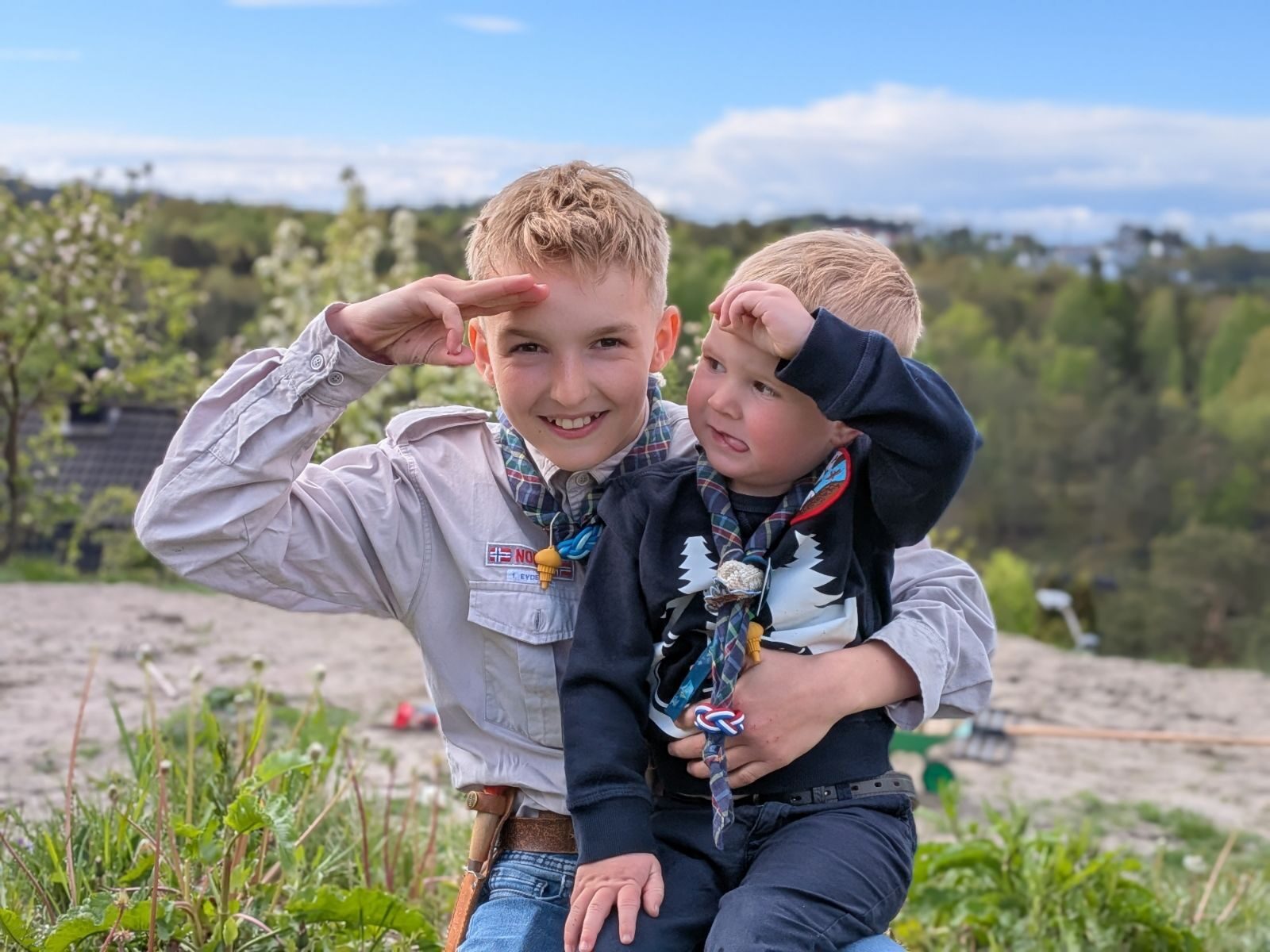 To unge gutter i norske speideruniformer hilser utendørs på en solskinnsdag med grønne trær bak seg.