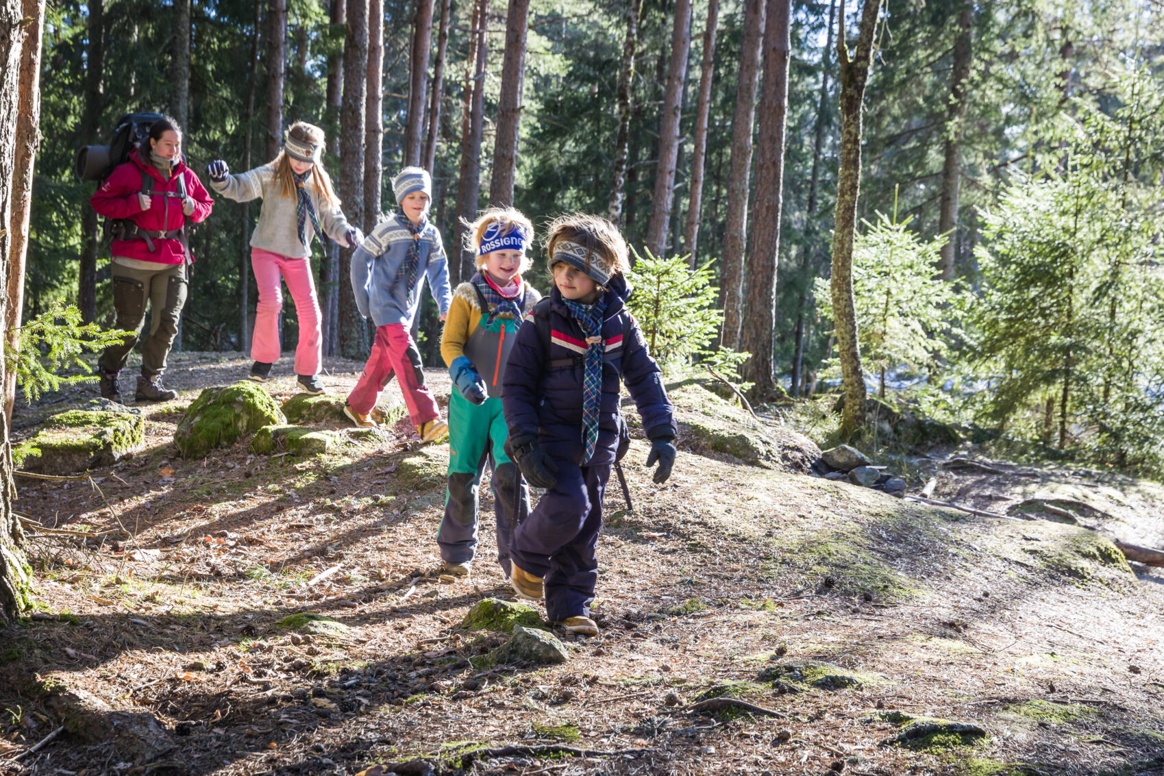 Gruppe barn vandrer gjennom en furuskog, iført fargerike utendørsklær og ryggsekker på en solskinnsdag.