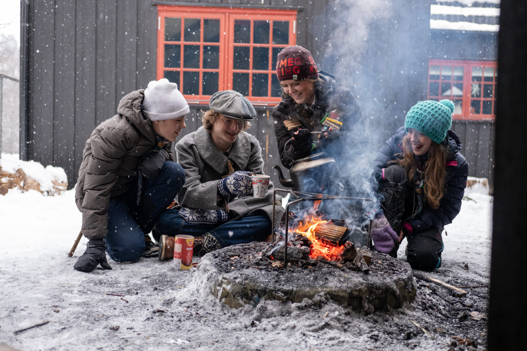 Gruppe mennesker sitter rundt et bål i snødekte forhold, varmer seg og lager mat utendørs.