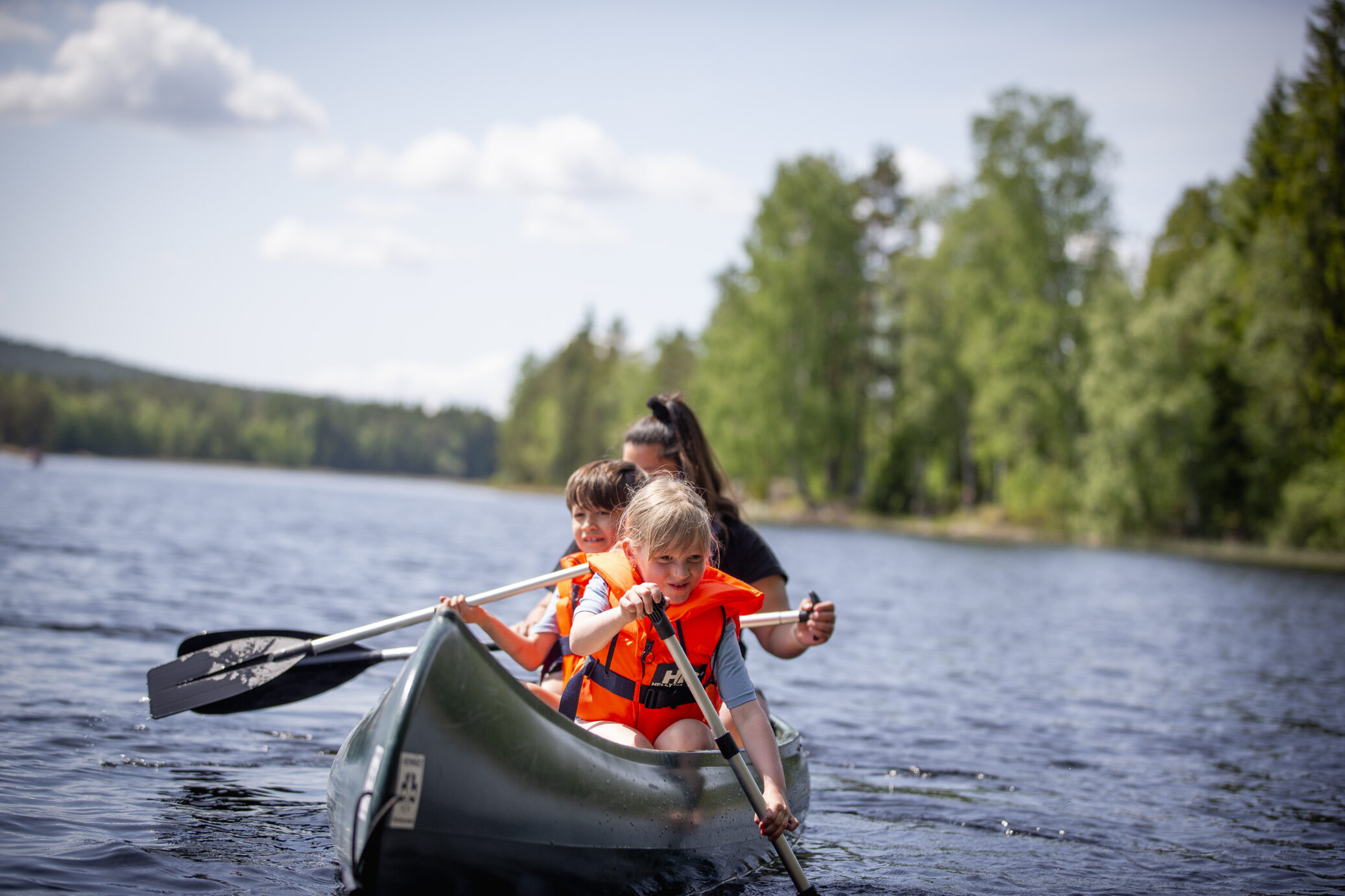 Familiepadling i kano på en innsjø omgitt av skog. To barn i oransje redningsvester padler mens en voksen guider bakfra.