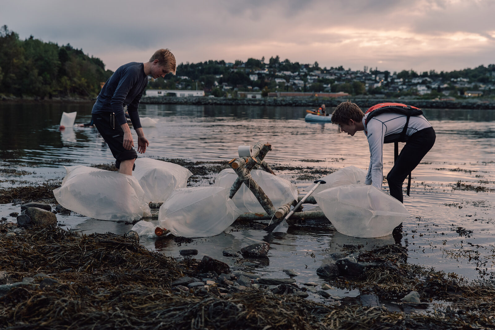 To speidere bygger en flåte av stokker og plastposer ved vannkanten i skumringen