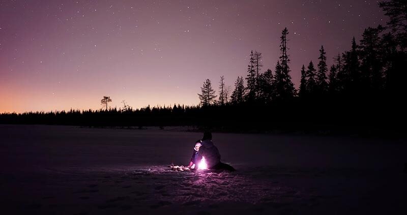 Person sitter ved et lite bål i vinterlandskap under stjernehimmel
