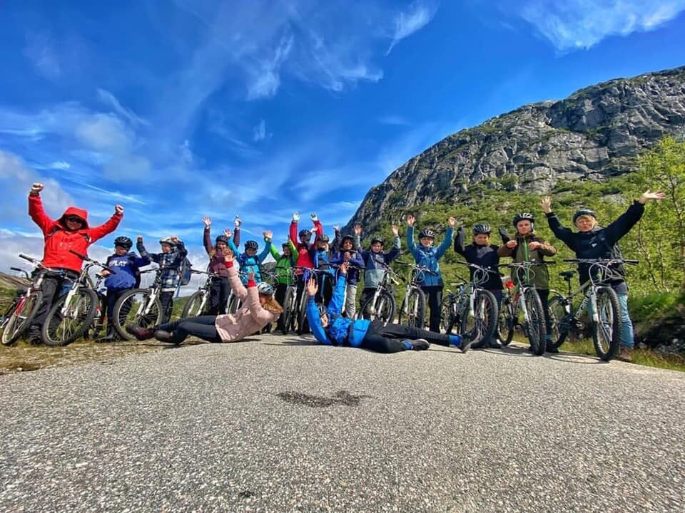 Gruppe speidere med sykler på en vei i fjellandskap, alle jubler med hendene i været